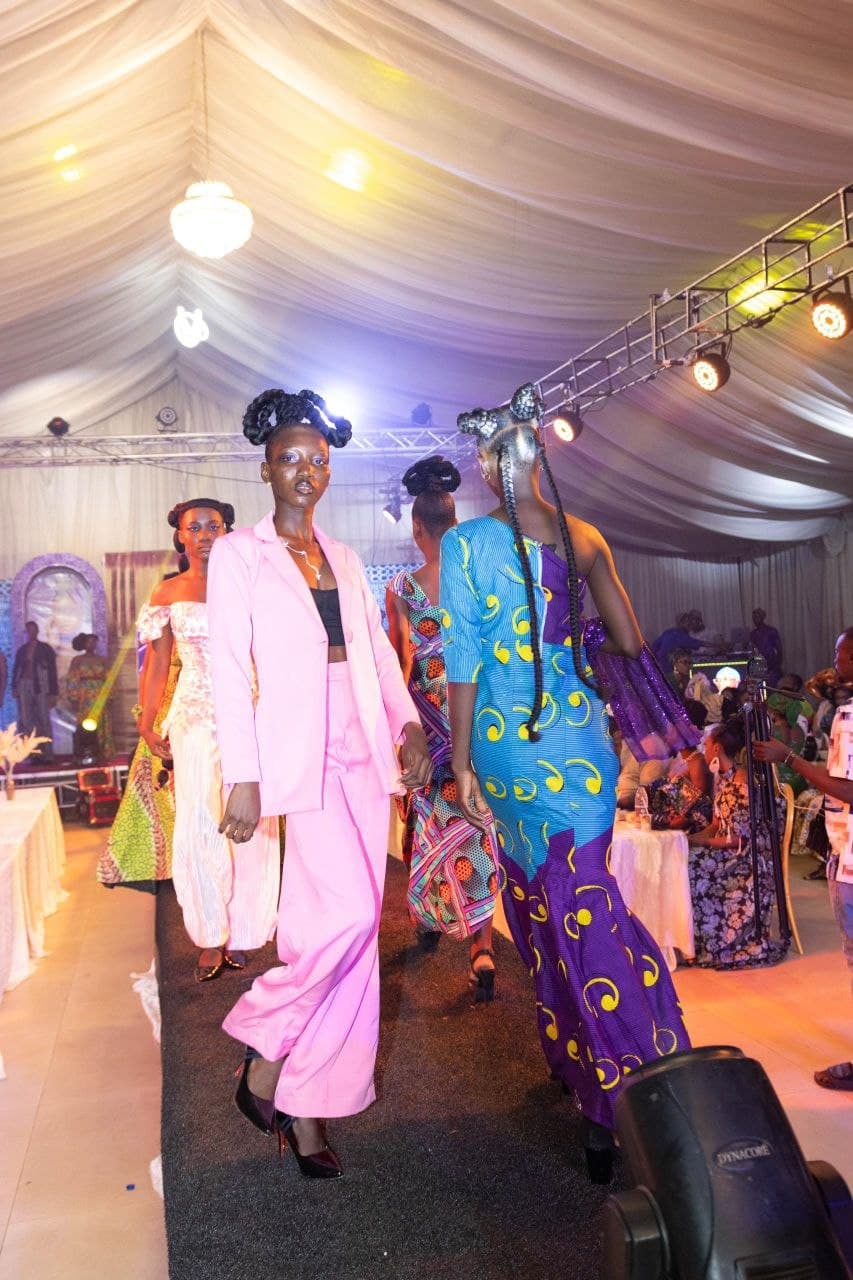 Models walk a runway in a pink suit and patterned dresses under bright stage lights.