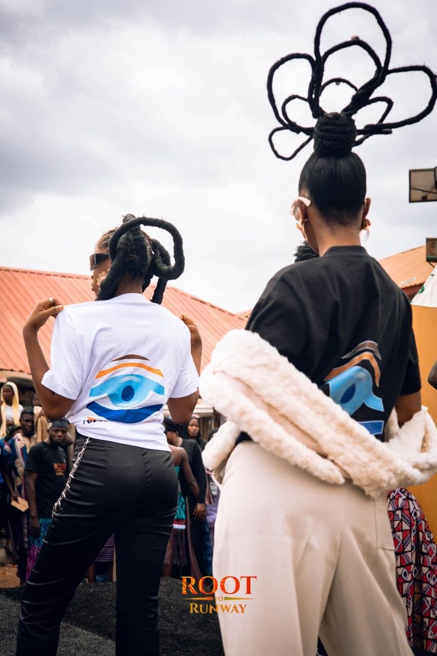 Models with elaborate sculptural hairstyles wear t-shirts with eye logos at an outdoor event.