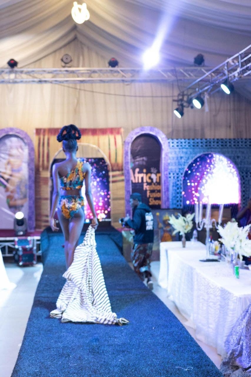 Model in patterned swimwear walks a blue runway, dragging a long, striped fabric behind her.