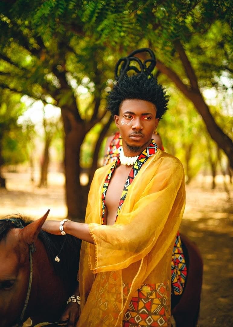 Man with face paint and yellow robe wearing shell jewelry standing beside a horse.