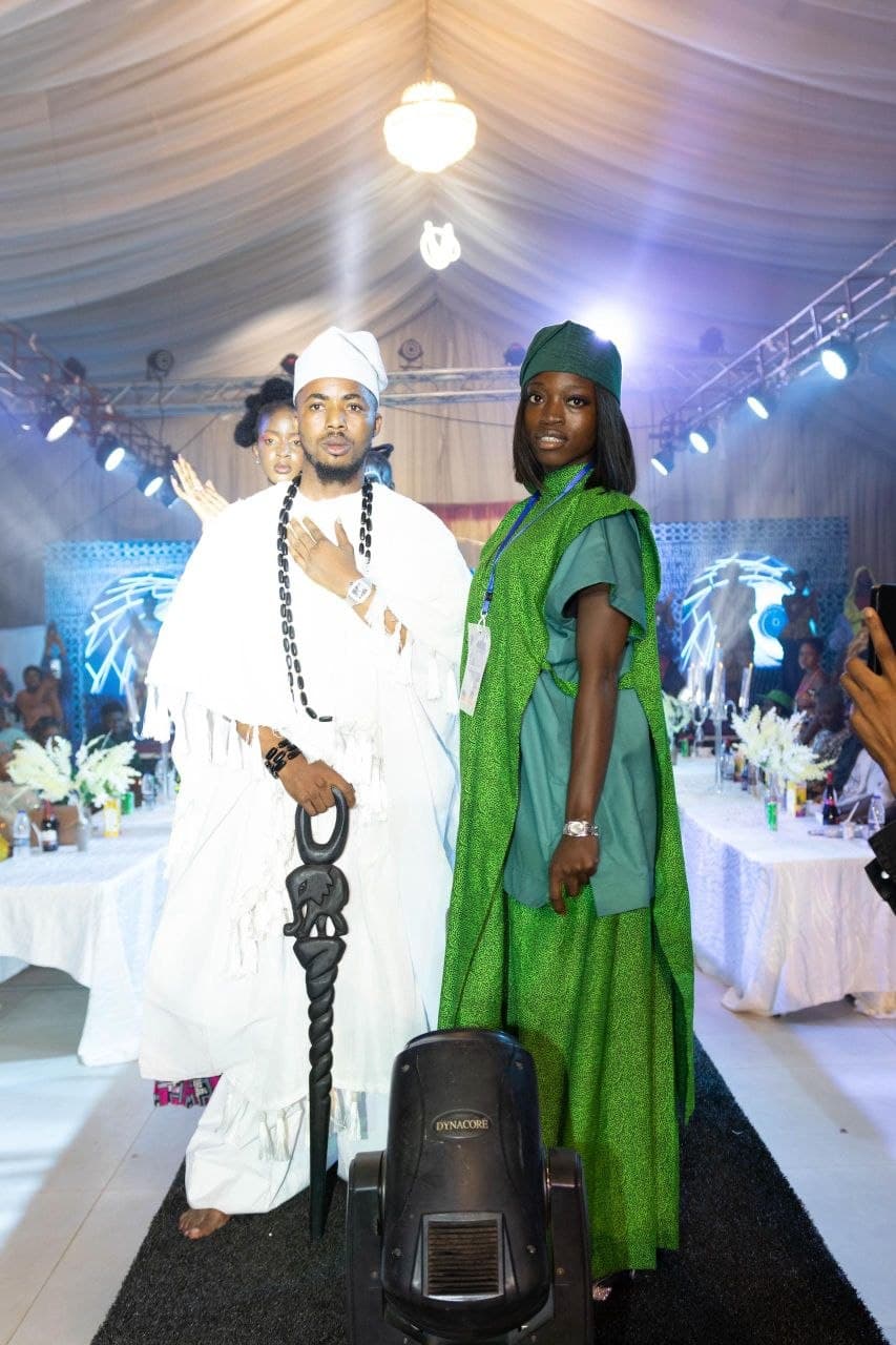 Man in white and woman in green traditional Nigerian attire pose on a fashion runway.