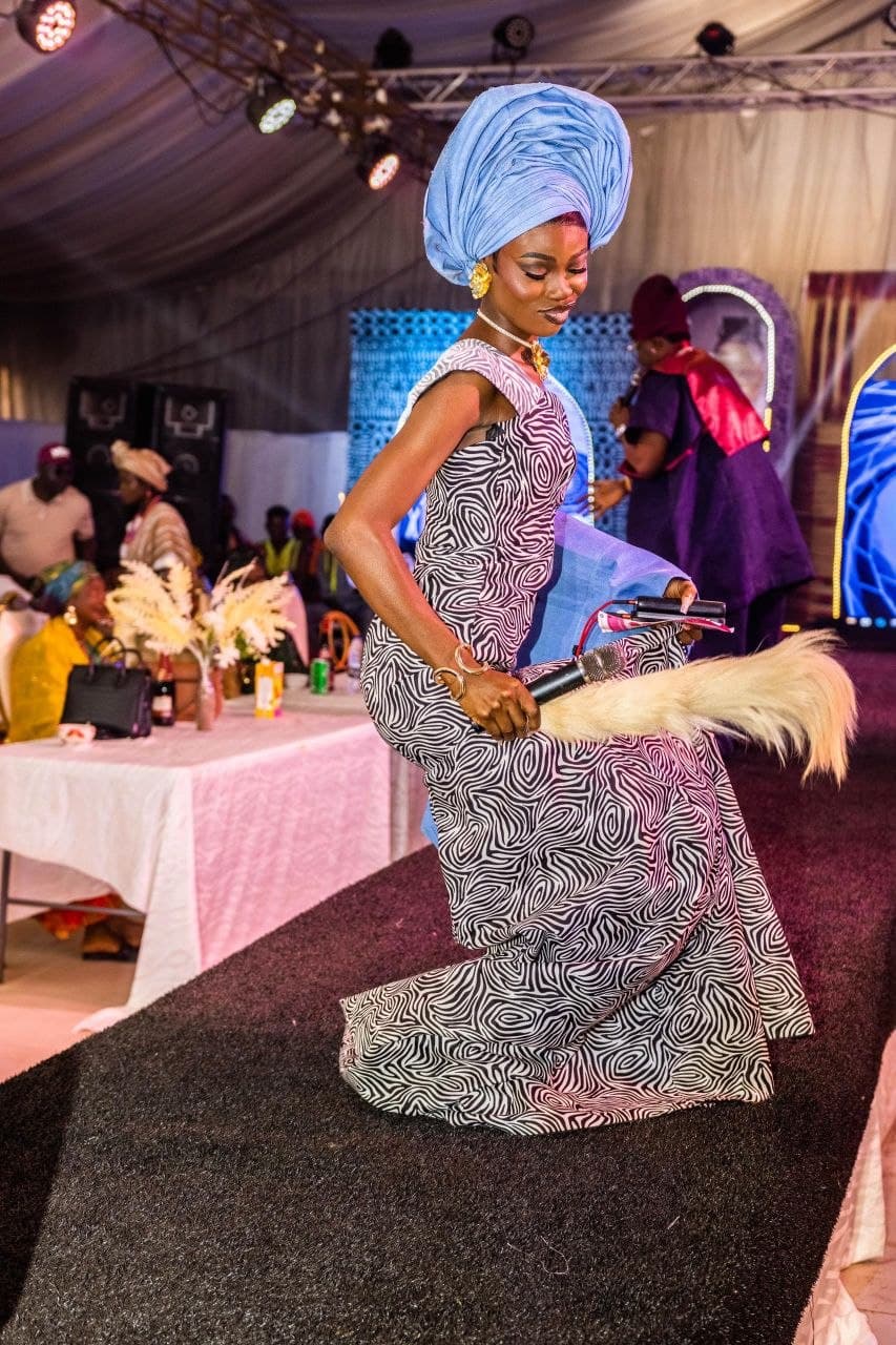 Woman in a patterned dress and blue gele holding a whisk on a runway.