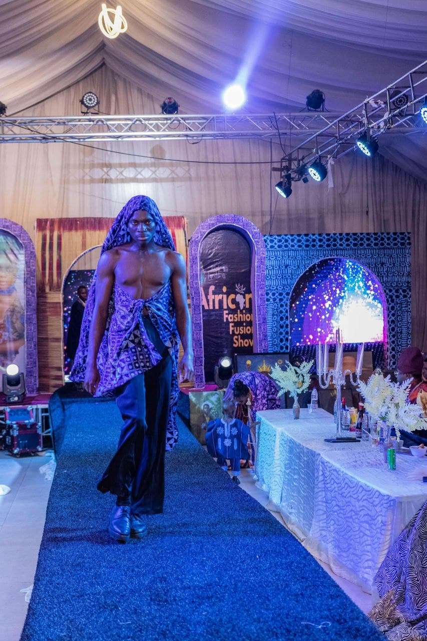 Male model walks a blue runway wearing a purple patterned headpiece and draped fabric.