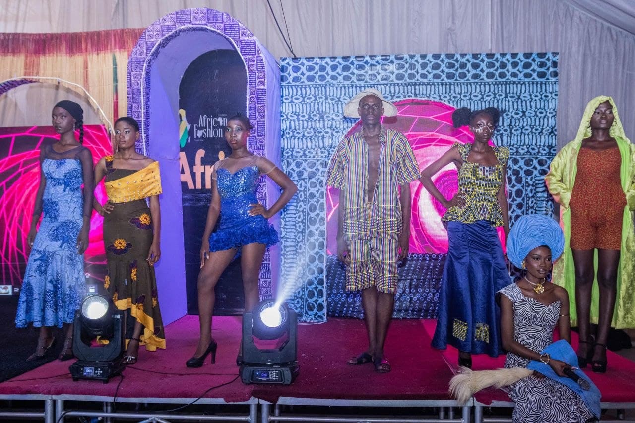 Models wearing vibrant African-inspired clothing pose on a stage during a fashion show event.