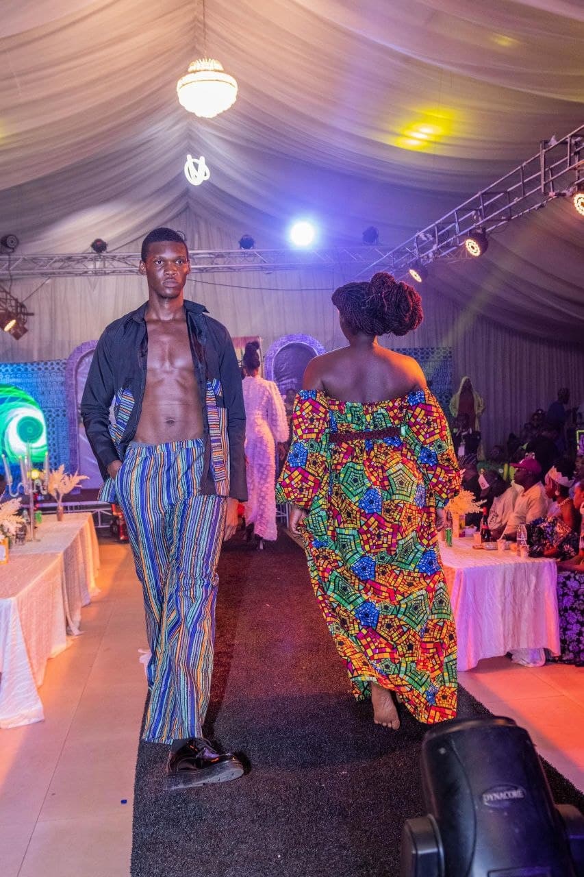 Male and female models walk a runway showcasing vibrant, patterned clothing in a tented venue.